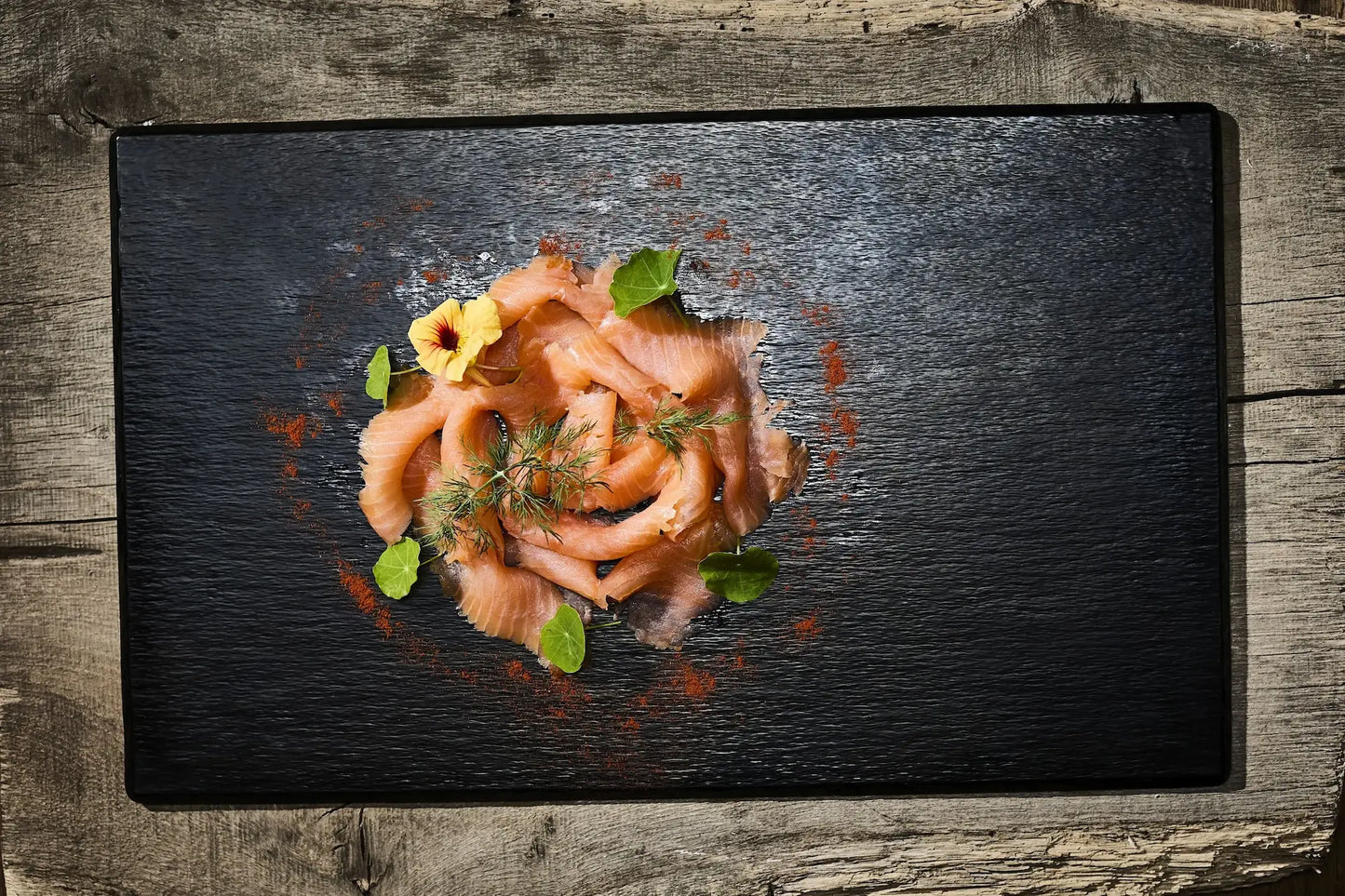 100 g Räucherlachsscheiben auf einer schwarzen Platte mit einer gelben Blumen und grünen Blättern