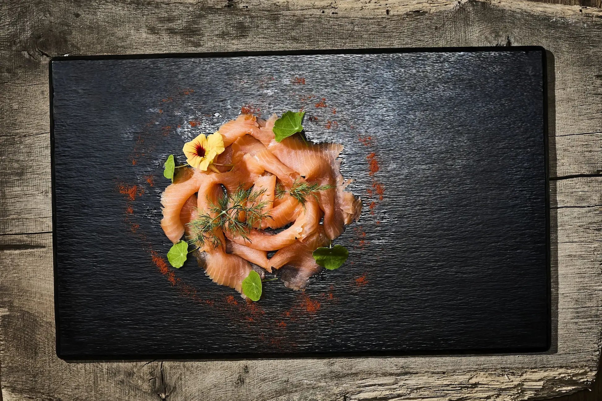 100 g Räucherlachsscheiben auf einer schwarzen Platte mit einer gelben Blumen und grünen Blättern 